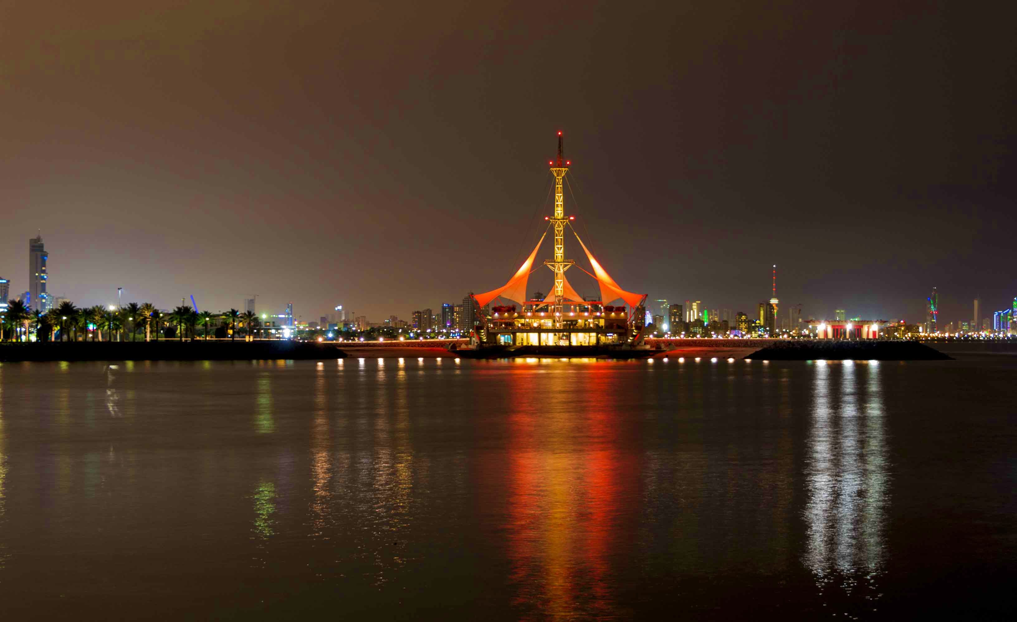 Illuminated traditional Omani pavilion structure reflected in waterway at night with city skyline and palm trees in backgroun